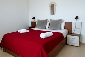 A neatly made bed with a red blanket, two pillows, and two rolled towels. There is a bedside table with drawers and a wall-mounted lamp. - Cosy apartment with a great sea view! Public Parking near the app! 60m2! (Sarandë)