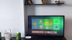Un soggiorno moderno con una TV a schermo piatto, un cartello decorativo che elenca città di partenza e una mensola minimalista con una pianta e una sveglia.