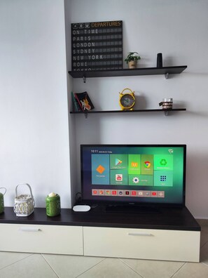 A modern living room with a flat-screen TV, a decorative sign listing departure cities, and a minimalist shelf with a plant and a clock. - Cosy apartment with a great sea view! Public Parking near the app! 60m2! (Sarandë)