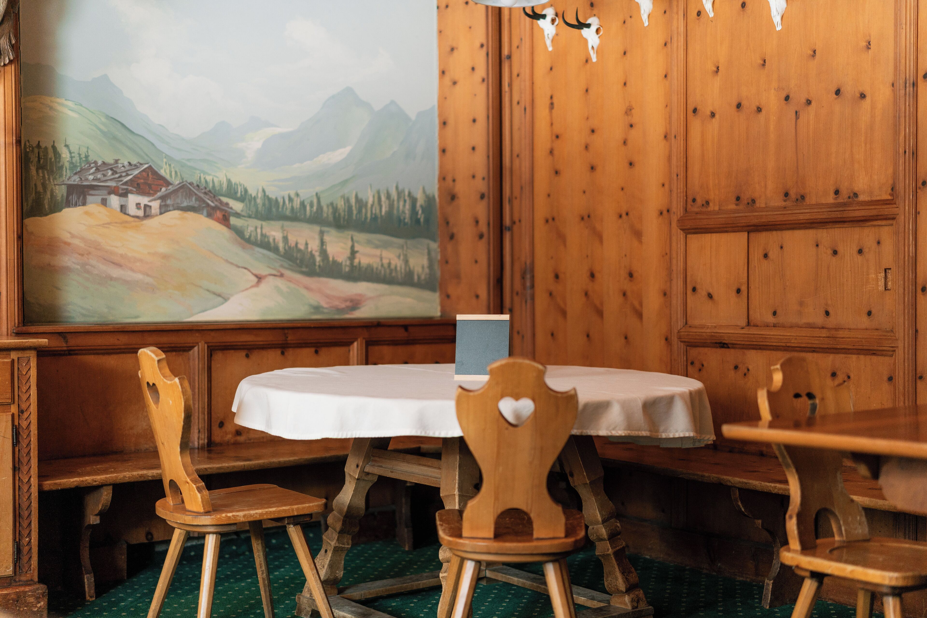 Uma mesa de jantar de madeira com toalha branca, rodeada por cadeiras de madeira com detalhes em formato de coração. Uma pintura grande de uma paisagem de montanha é visível na parede atrás da mesa.