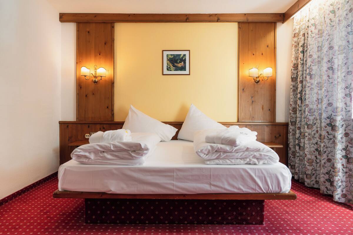 A hotel room with a wooden headboard, two wall sconces, a framed picture, and a red patterned carpet.