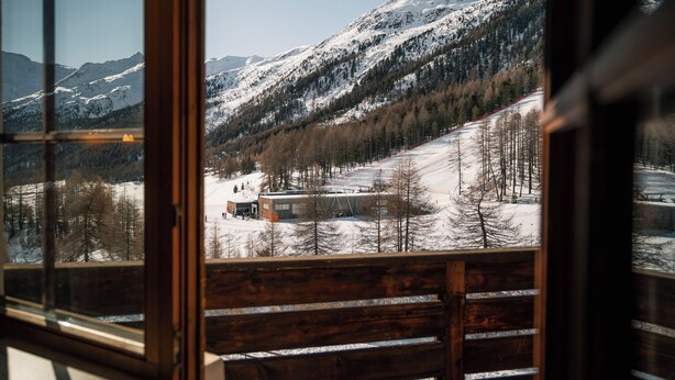Vista desde una ventana con marco de madera, sobre un paisaje montañoso cubierto de nieve con una estación de esquí y edificios.