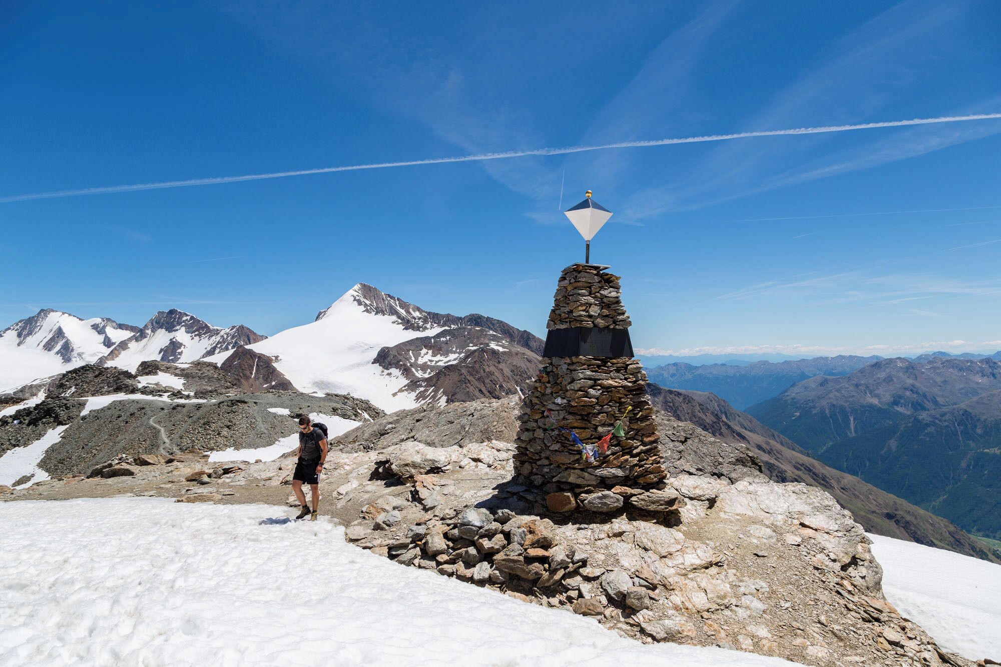 A stone cairn with a flag on a snowy mountain peak, surrounded by rugged terrain and distant mountains.