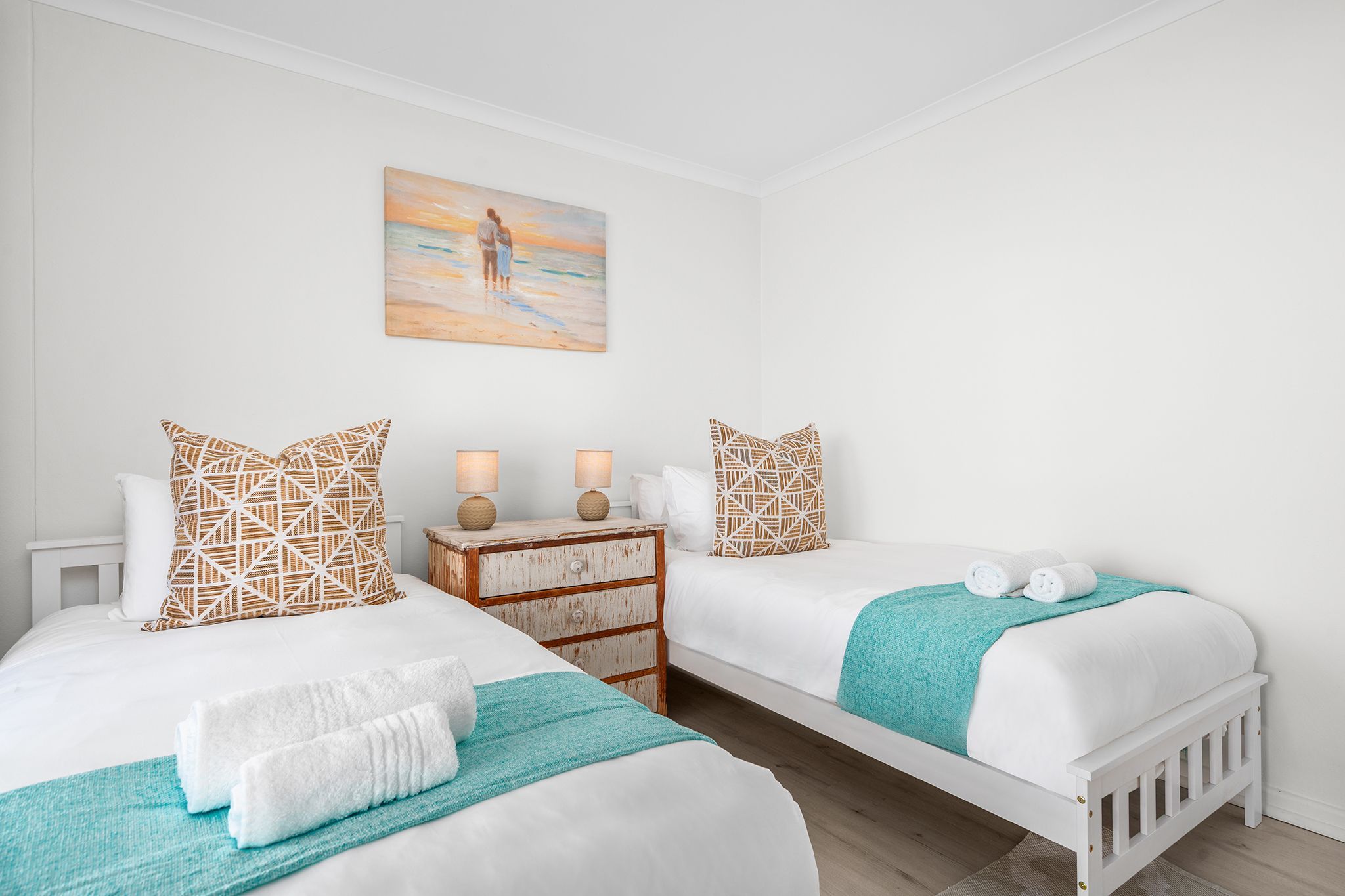 Un dormitorio con dos camas, una mesita de noche, un cuadro y una manta color aguamarina.