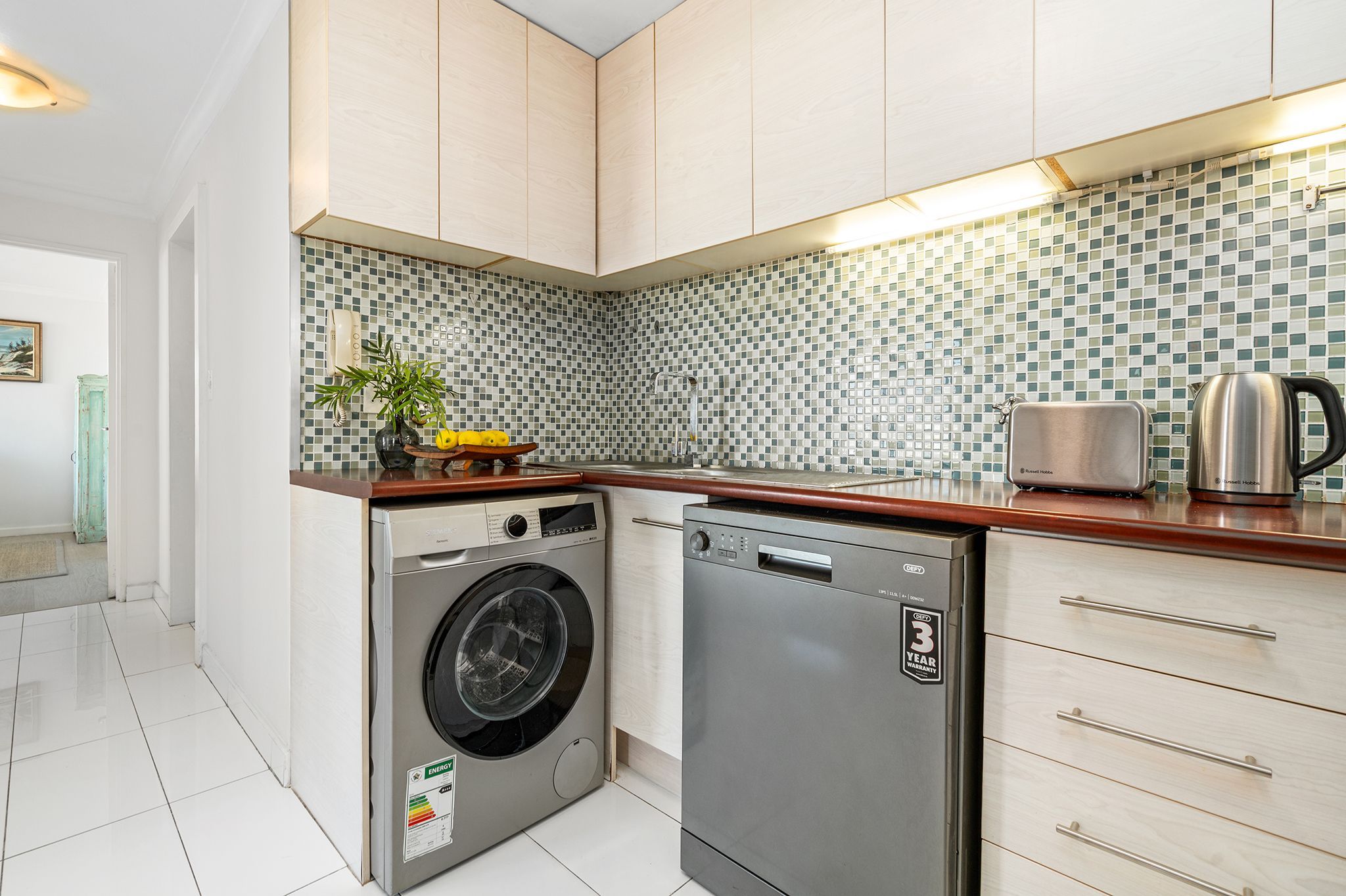 A compact kitchen with a washing machine, a dishwasher, a tiled backsplash, and wooden cabinets.