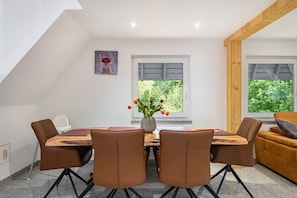 A dining area with a wooden table, four brown chairs, a vase with flowers, and two windows with a view of greenery.