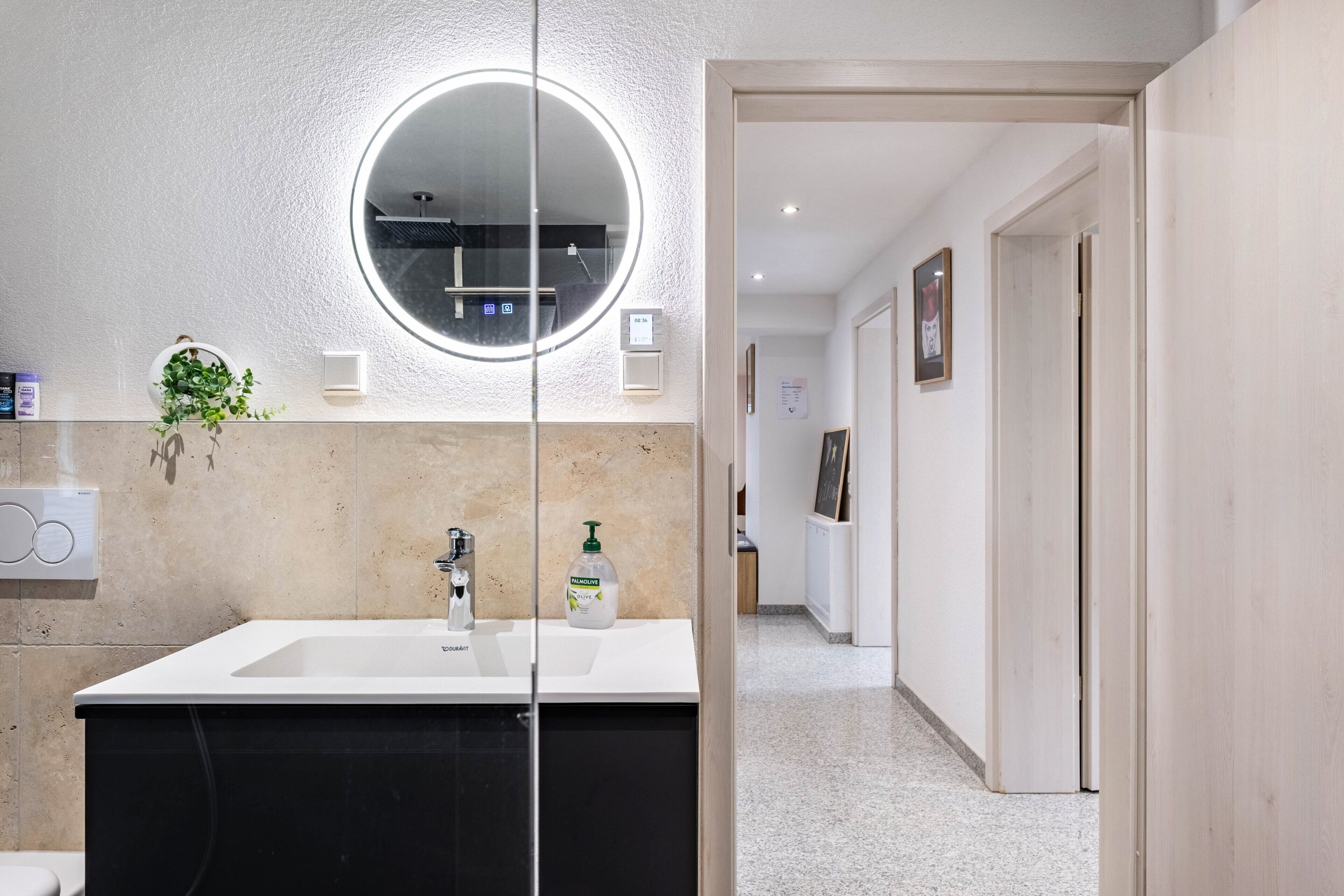 Un baño moderno con un lavabo blanco, un espejo redondo, una planta y un dispensador de jabón.