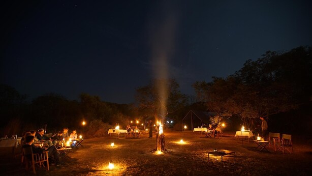Ein Campingplatz bei Nacht, mit Tischen und Stühlen um ein zentrales Lagerfeuer angeordnet.