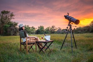 A person sitting on a chair in a grassy field with a telescope on a tripod. - Mahoora Tented Safari Camp Wilpattu (Pahala Maragahawewa)