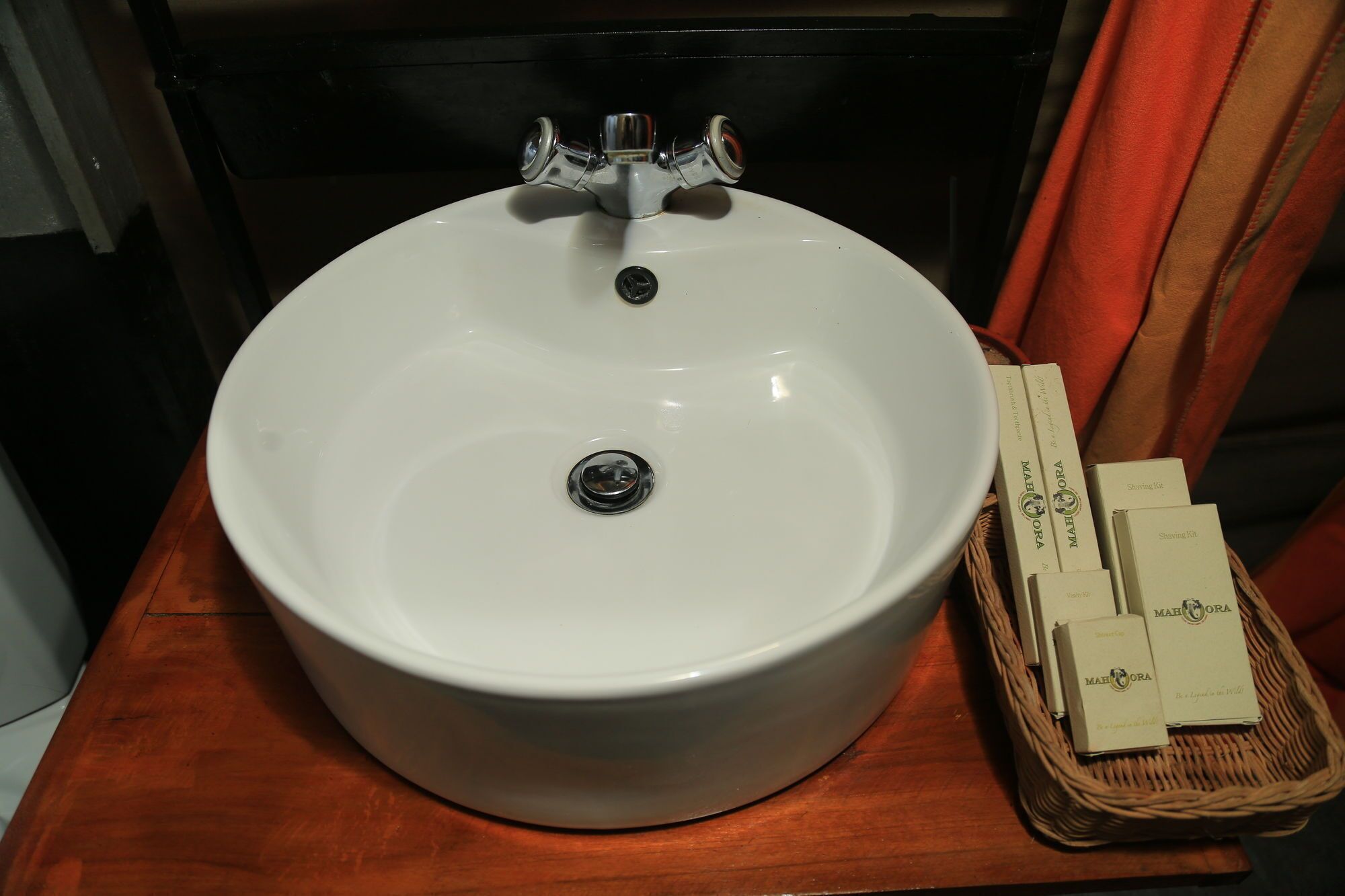 A white bathroom sink with a faucet, a mirror, and a wicker basket containing Mahora shaving kits.
