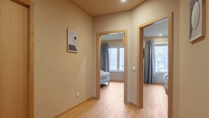 Een hal met houten vloeren, een deur links en een bed met raam en gordijnen op de achtergrond.