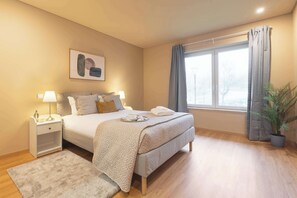 Une chambre bien rangée, avec un lit, des tables de chevet, une fenêtre avec des rideaux et une plante en pot.