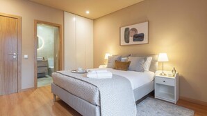 Een slaapkamer met een bed, nachtkastjes, een deur en een zichtbare badkamer door een geopende deur.