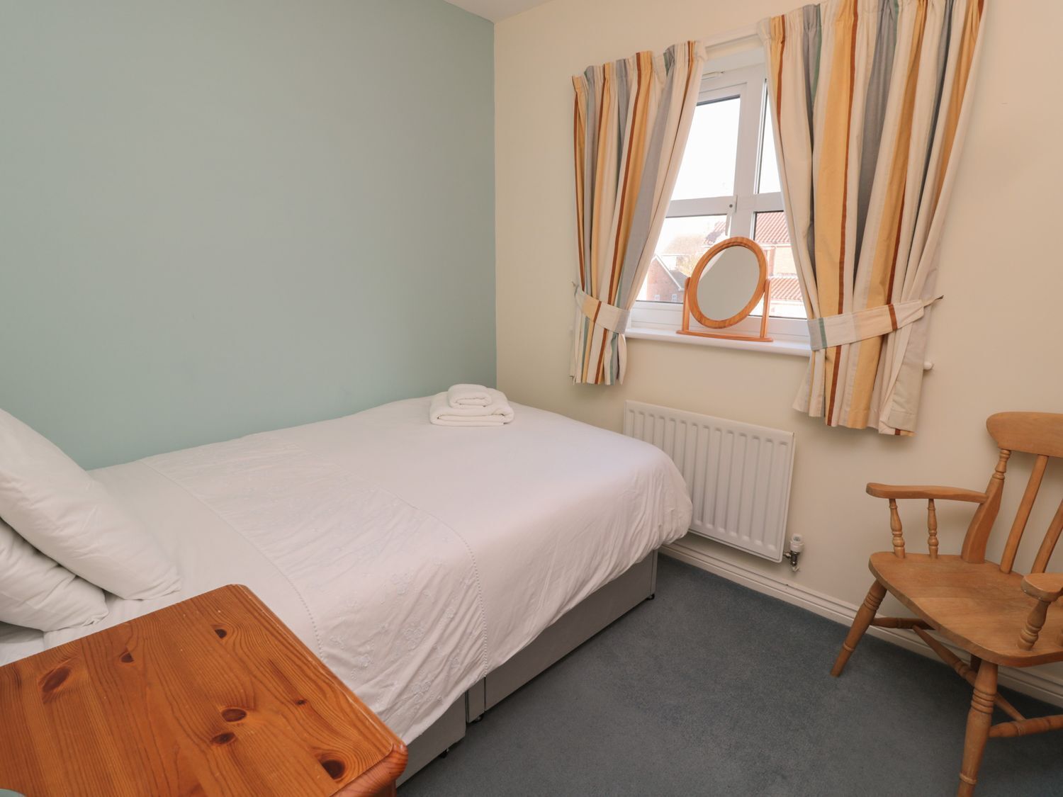 Quarto com uma cama, uma cadeira de madeira, uma pequena mesa de madeira, um radiador e uma janela com cortinas.