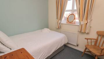 Quarto com uma cama, uma cadeira de madeira, uma pequena mesa de madeira, um radiador e uma janela com cortinas.