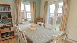 A dining area with a polka dot tablecloth, white chairs, a wooden bookshelf, and a window with curtains. - Sandy Cove (Chathill)