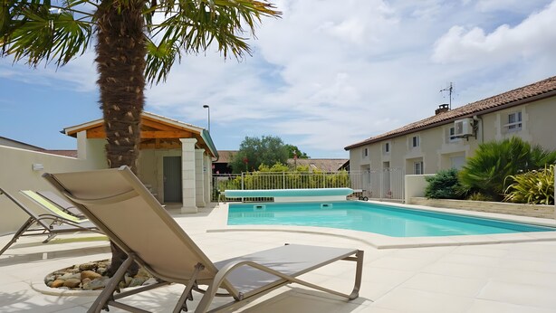 A swimming pool with lounge chairs, a palm tree, and a building in the background. - Family Guest Room Anis (Semussac)