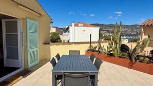 Een terras met een tafel, stoelen, een schuifdeur en uitzicht op gebouwen en bergen.