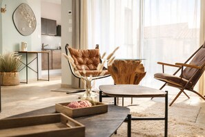 Uma sala de estar moderna com sofá, duas poltronas, mesa de centro e mesa lateral de madeira. Há uma planta em vaso e um espelho decorativo na parede.