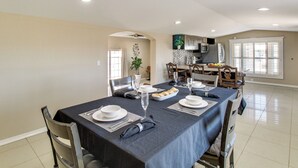 Un coin repas avec une longue table dressée pour quatre personnes, un ventilateur de plafond et une cuisine en arrière-plan.