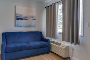Una habitación con un sofá azul, un cuadro en la pared, una ventana con cortinas y un aire acondicionado instalado en la pared.