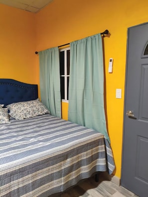 Une pièce avec un lit, une fenêtre avec des rideaux, une porte et un interrupteur au mur.