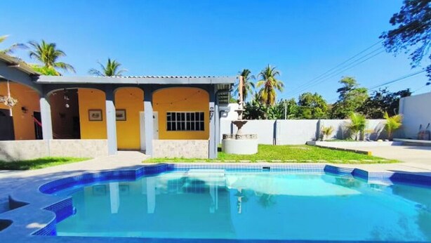 Une piscine avec un bord carrelé, un mur blanc orné d’une fontaine et un bâtiment aux murs jaunes et blancs, surmonté d’un toit en tuiles.