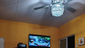 Une pièce avec un téléviseur fixé au mur, un haut-parleur sur un trépied, une table ronde agrémentée d’un objet décoratif et un ventilateur de plafond.