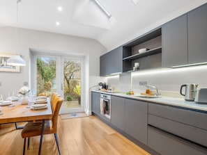 A modern kitchen with grey cabinets, a wooden dining table, and a view of a garden through a glass door. - Market Street Apartment Clifden Town (Clifden)