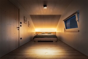 Ein Schlafzimmer mit Holzboden, einem Bett mit Kopfteil, einem Fenster mit Blick auf den Nachthimmel und einer Tür links.