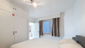 Un dormitori amb un llit, un armari, una finestra amb cortines i una porta que dona a una altra habitació.