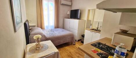 A compact hotel room with a bed, a small table with a vase, a kitchenette with a stove, and a TV on a cabinet.