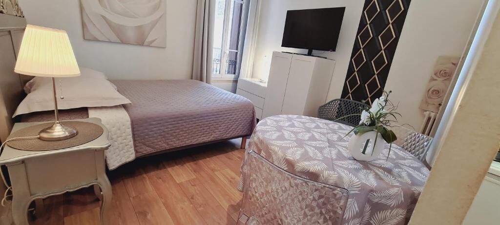 Une chambre à coucher avec un lit, une table de chevet avec une lampe, un téléviseur posé sur un meuble, une petite table avec un vase et une fenêtre avec des rideaux.