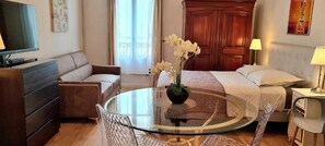 Une chambre d’hôtel avec un lit, une table ronde en verre sur laquelle est posé un vase de fleurs, un canapé, un téléviseur et une armoire en bois.