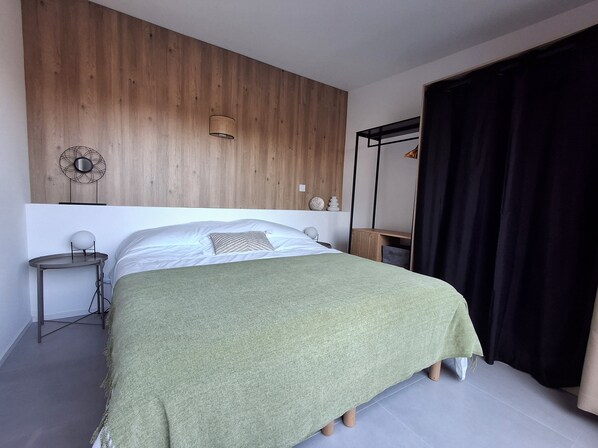 Quarto com cabeceira de madeira, cama com manta verde, mesa de cabeceira com luminária e cortina preta.