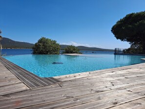 Um deck de madeira leva a uma piscina azul límpida, com vista para o mar e colinas ao longe.