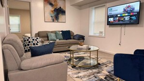 Ein Wohnzimmer mit einem beigen Sofa, einem blauen Sessel, einem Glastisch und einem an der Wand montierten Fernseher.