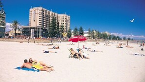 Une plage où des gens se prélassent au soleil, un parasol à l’effigie de Coca-Cola et, au loin, un grand immeuble d’hôtel.