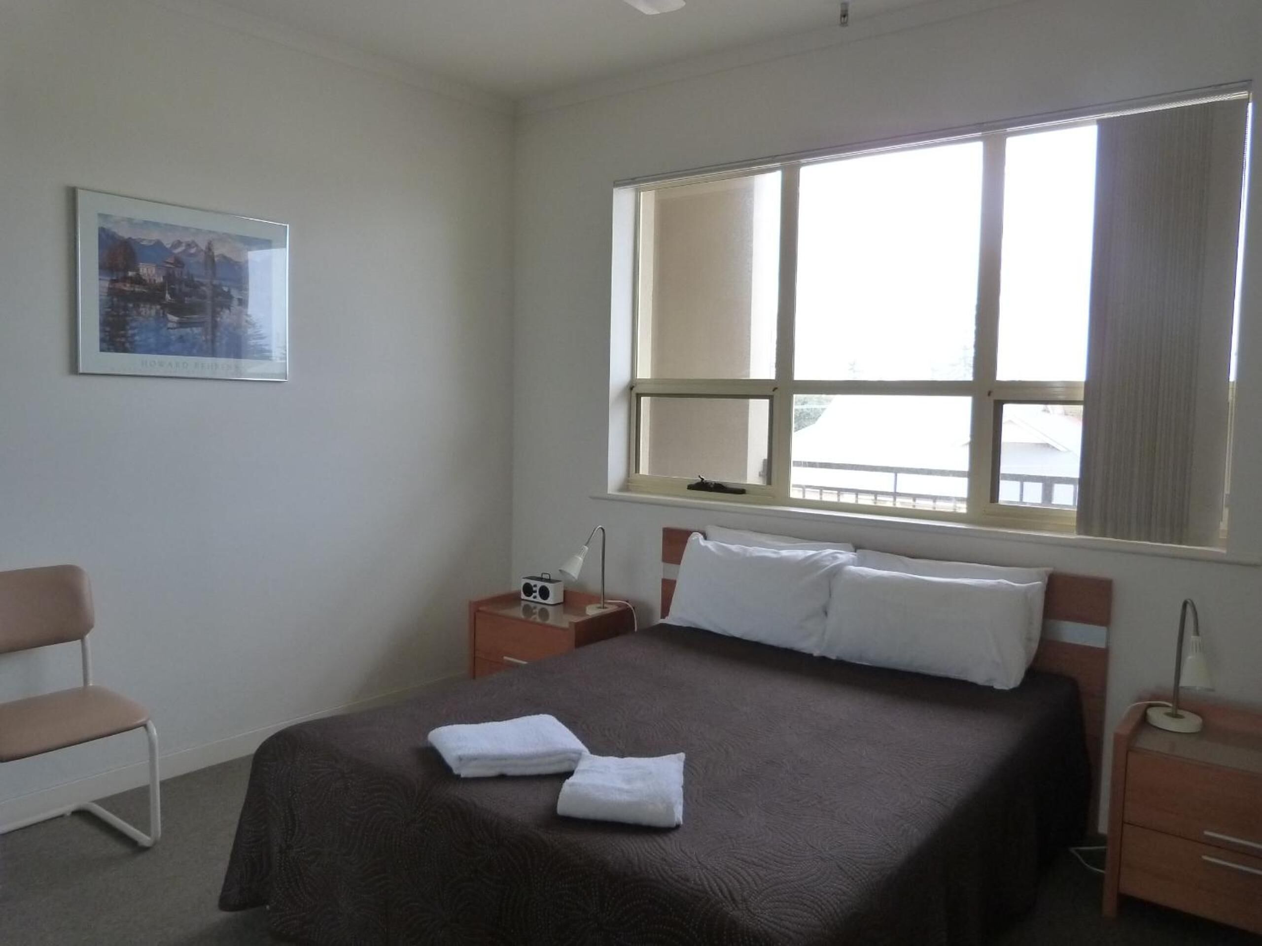 Una camera d'albergo con un letto, una sedia, un comodino con lampada e una finestra con tende.