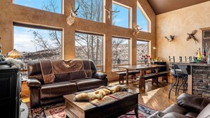 A spacious living room with a leather sofa, a coffee table, and a fireplace. Large windows offer views of the outdoors.