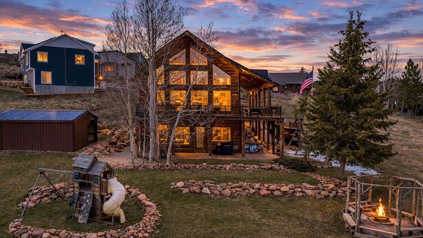 A large log cabin with a balcony, surrounded by a stone wall and a wooden playset.