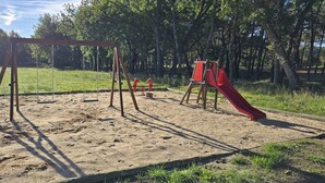 Leikkikenttä, jossa on keinut ja punainen liukumäki, ja taustalla näkyy puita ja ruohikkoista aluetta.