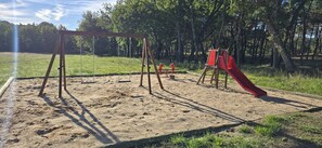A playground with swings and a red slide in a grassy area with trees in the background. - ZEcamping 3* piscine mobil-home clim 35m2 6 pers (Quinson)