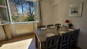 A dining room with a wooden table set for six, a radiator, a window with a view of trees, a red lamp, and a framed picture on the wall. - ZEcamping 3* piscine chiens acceptés locatif 43m2 4 pers (Quinson)