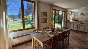 A dining area with a table set for four, a kitchen with a refrigerator, and a view of a garden through large windows. - ZEcamping 3* piscine pet friendly locatif 90m2 6 pers (Quinson)