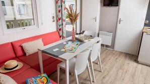 Un espacio habitable compacto con un sofá rojo, una pequeña mesa de comedor con sillas, un televisor montado en la pared y una ventana con persianas.