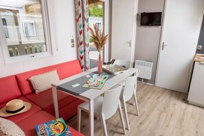 Ein kompakter Wohnbereich mit einem roten Sofa, einem kleinen Esstisch mit Stühlen, einem an der Wand befestigten Fernseher und einem Fenster mit Jalousien.