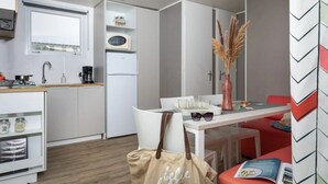 Una cocina compacta con armarios blancos, un microondas y un refrigerador. Hay un comedor con una mesa, sillas y un jarrón con plantas secas. Un bolso con texto está en el suelo.