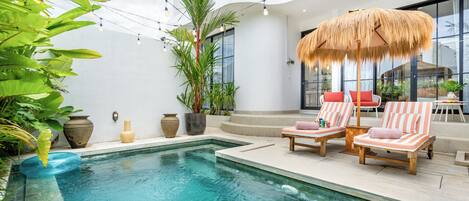 A pool area with lounge chairs and a thatched umbrella, surrounded by potted plants and a modern building with large windows.