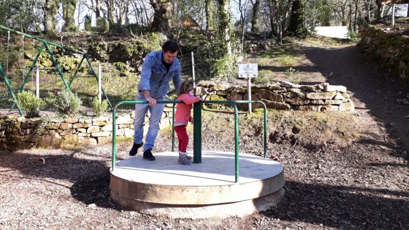 A man and a child on a circular platform with a metal railing, surrounded by a natural setting with trees and a stone wall.
